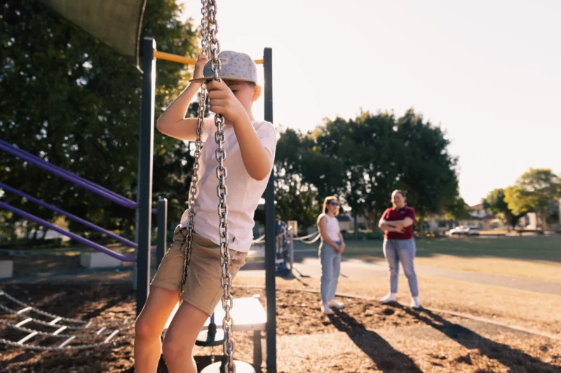 Child playing on playground while his mother and Anglicare worker chat about Anglicare' Child Social and Emotional Wellbeing Programs.