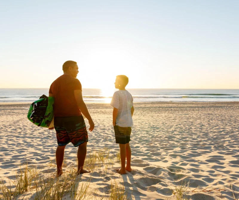 Father standing next to his son on the sand at a beach with a surfboard under his arm.