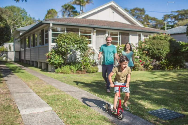Foster carers standing in front yard watching foster son on scooter.