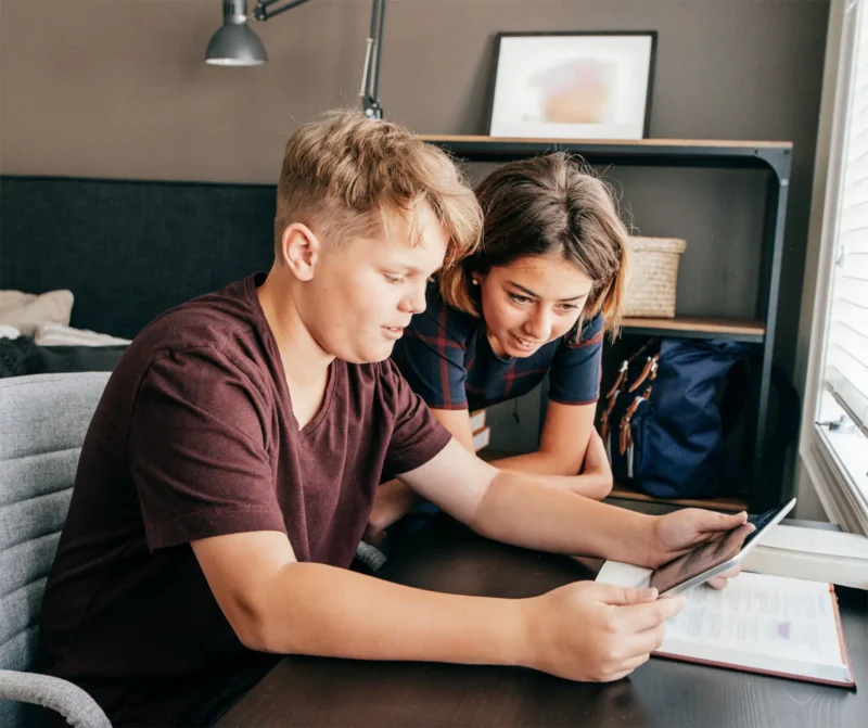 Two teenage foster children standing over a desk looking at an iPad.