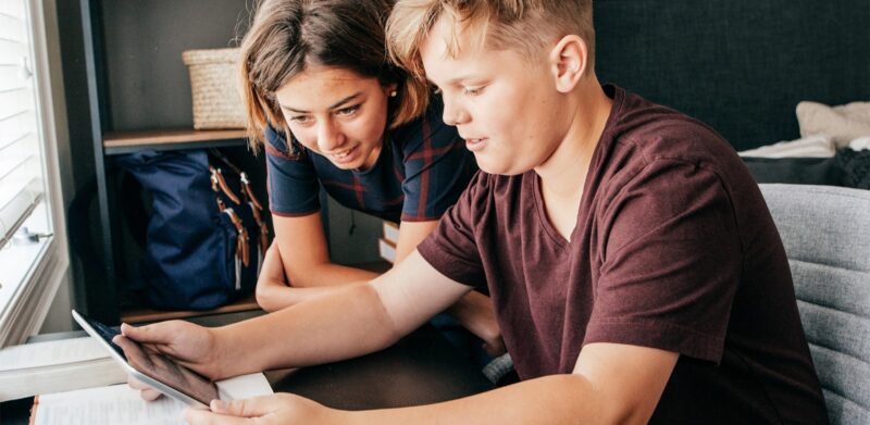 Image of two youth; young female and young male sitting at a desk looking at an iPad.