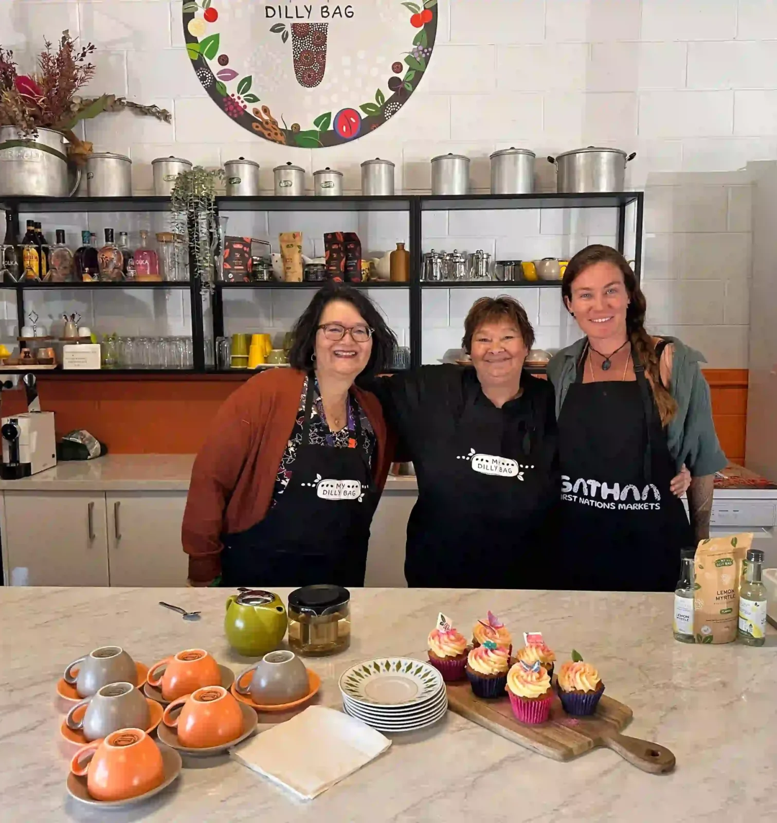 Three ladies standing behind a kitchen bench, smiling at the camera. There are cups and saucers on the bench and cupcakes too.